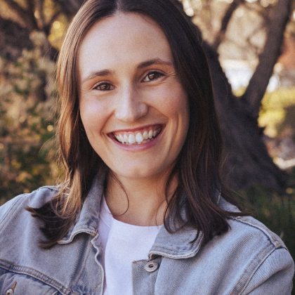 Dr Nicoli Cox smiles at the camera. She has long dark hair and is wearing a white top and pale denim jacket. there are trees in the background which are out of focus