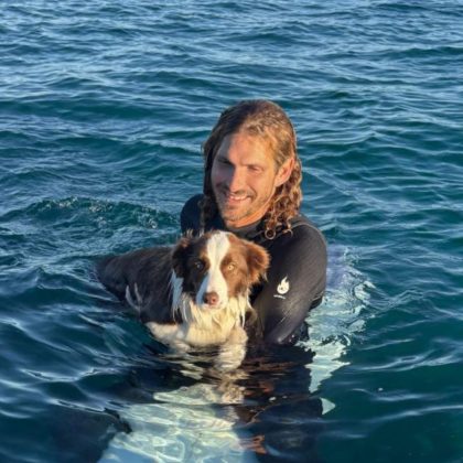 Dr Tim Archer smiles at the camera. He is on a surf board with his dog in the water.