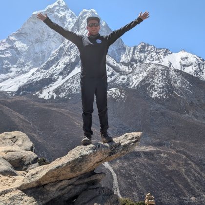 Dr Ben Holt standing on cliff edge with is hands in he air with mountains in the background